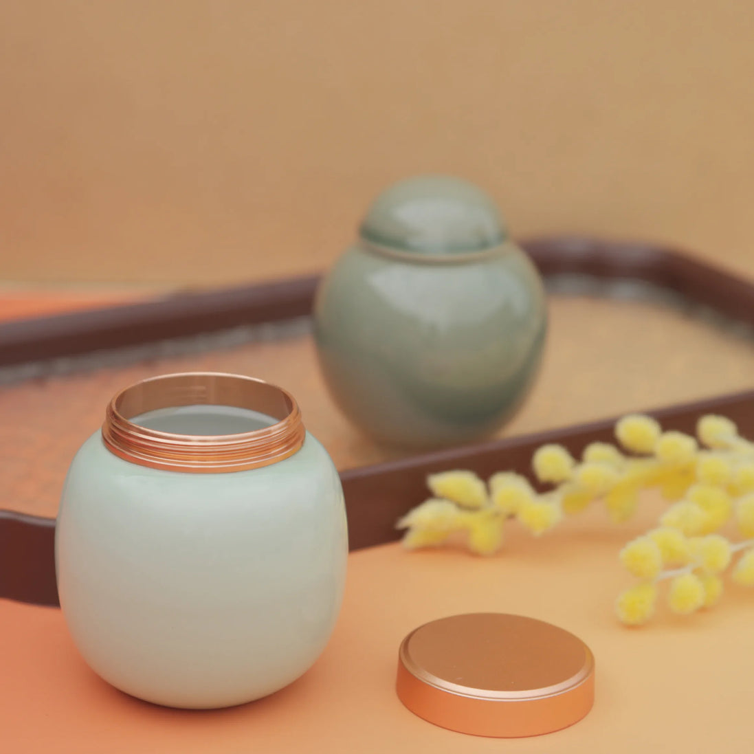 Lily jar with copper lid open on terracotta surface with yellow wattle flowers
