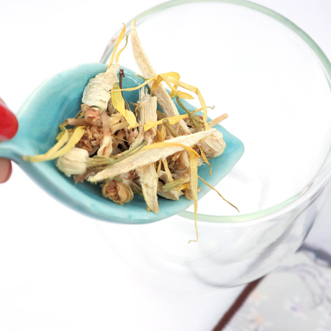 Hand holding turquoise measuring spoon filled with loose leaf herbal tea over glass jar