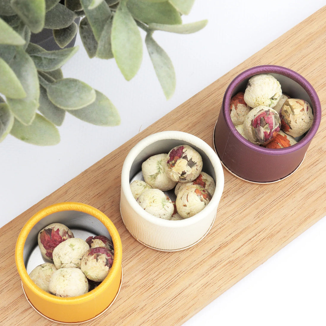 Three colourful tea tins with botanical tea balls on wooden serving board