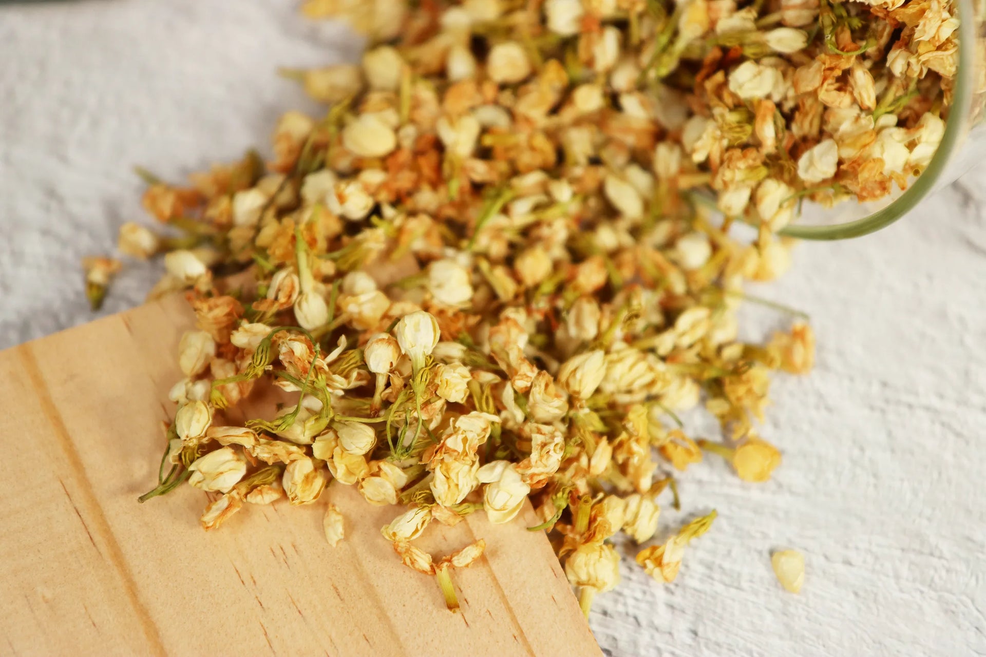 Golden dried jasmine flowers spilling from wooden spoon onto white marble surface with scattered petals