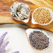 Three ceramic bowls containing dried herbs - gray sage leaves, golden sandalwood chips, and purple lavender buds