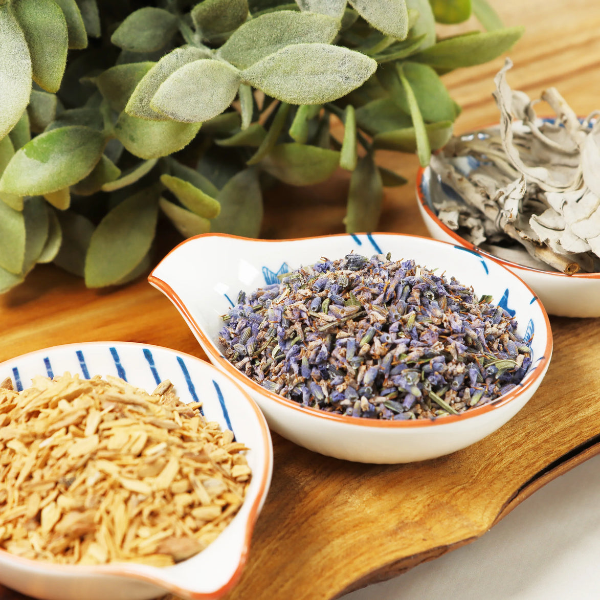Two ceramic bowls on wooden surface containing dried lavender blend and sandalwood chips with sage leaves nearby