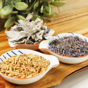 Three ceramic bowls containing dried sandalwood chips, lavender buds, and sage leaves on wooden tray with green plants