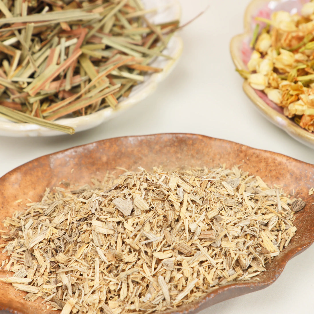 Dried agarwood chips, lemongrass, and jasmine flowers arranged in rustic ceramic dishes on white surface
