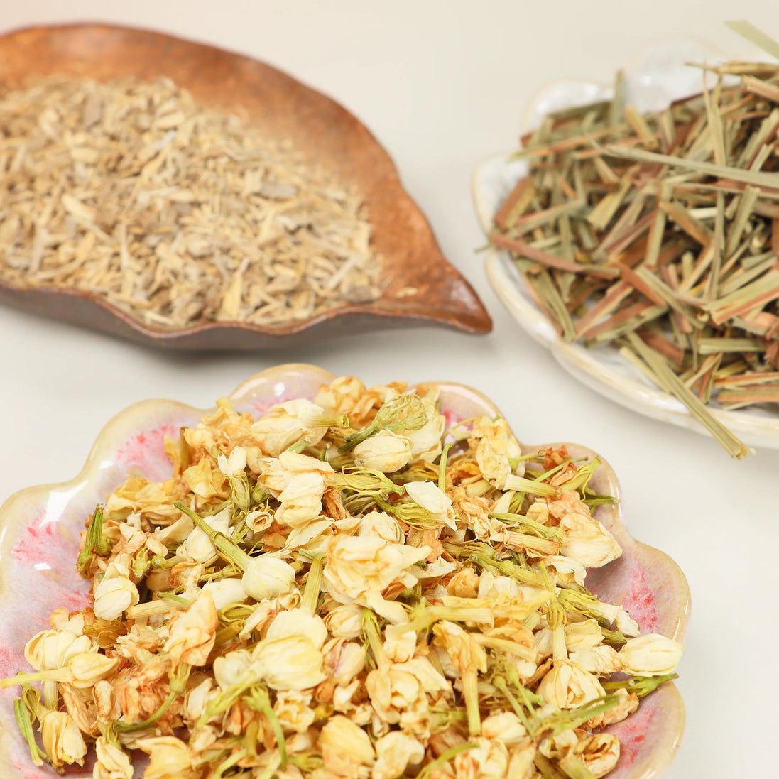 Three decorative dishes displaying dried agarwood chips, lemongrass strands, and jasmine flower blend on white surface