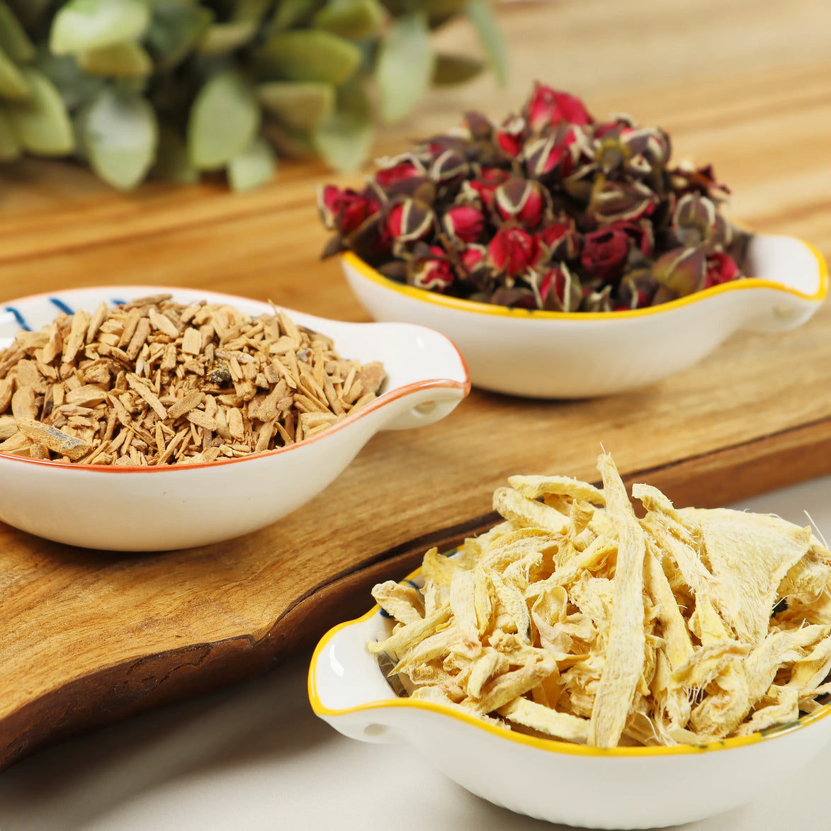 Two decorative bowls containing dried red roses and golden sandalwood chips on wooden cutting board