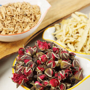 Bowls of dried rose petals, sandalwood chips, and ginger pieces arranged on wooden cutting board for aromatherapy blend