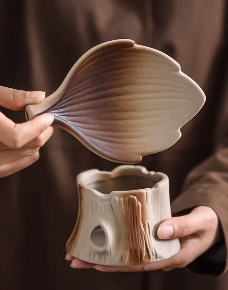 Hands holding leaf-shaped lid and tree stump ceramic aromatherapy burner in cream and brown tones