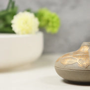 Grey ceramic incense burner with gold lotus petal pattern sits on white surface next to white vase with flowers