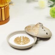 Grey ceramic incense burner with gold lotus petal design and ventilation holes, lid partially open showing interior