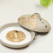 Grey ceramic incense burner with gold lotus petal design, lid open showing white interior with geometric pattern