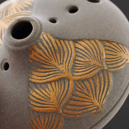 Grey ceramic incense burner with gold palm leaf pattern and ventilation holes on dark background