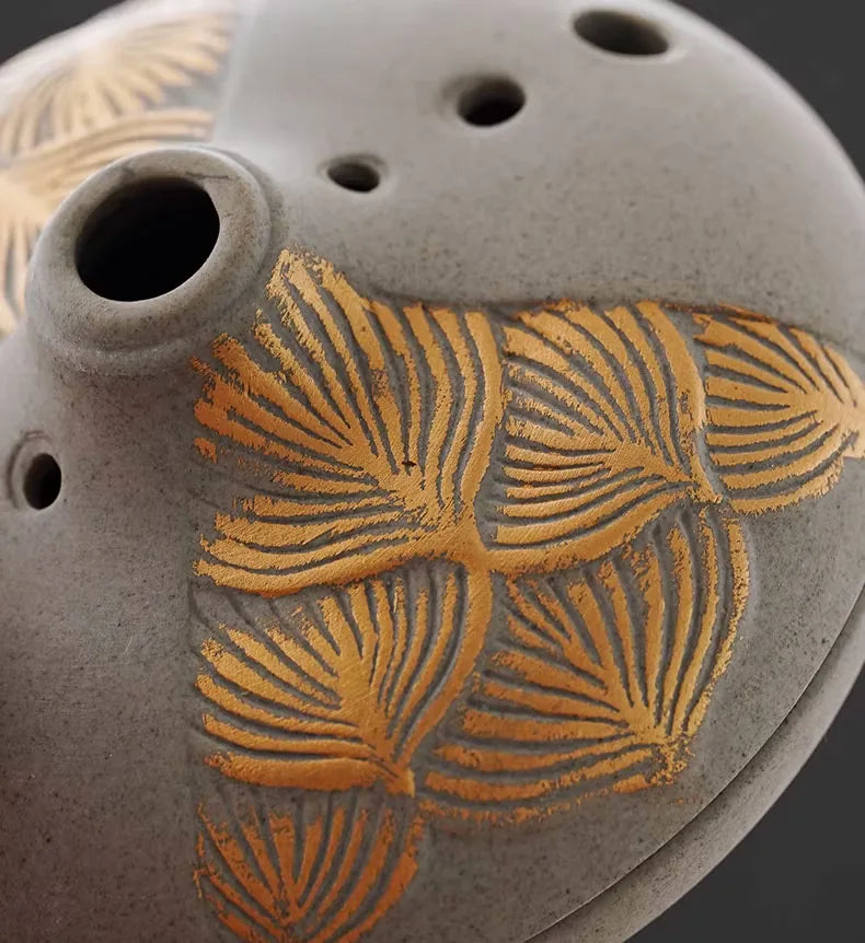 Grey ceramic incense burner with gold palm leaf pattern and ventilation holes on dark background