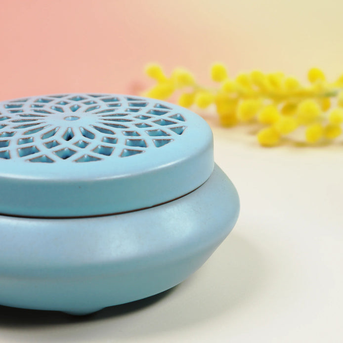 Powder blue ceramic incense burner with geometric lattice lid and yellow flowers in background