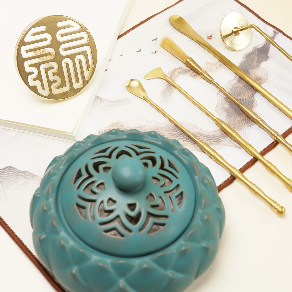 Turquoise ceramic incense holder with carved patterns, gold prosperity seal, and brass ritual tools on white surface