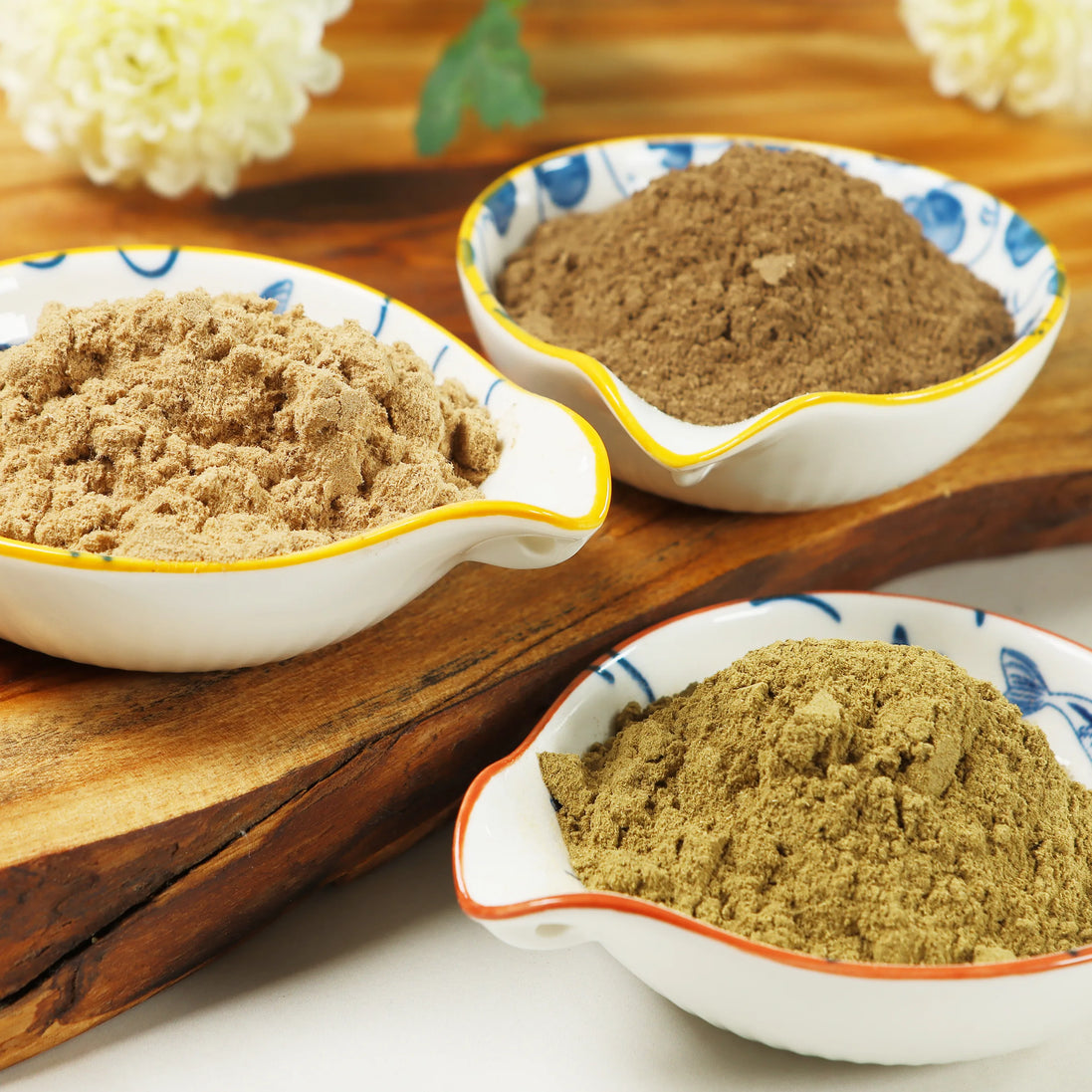 Three ceramic bowls containing natural incense powder blends in beige, brown, and olive green colors on wooden surface