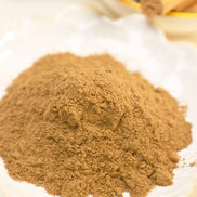 Fine golden-brown incense powder mounded on white plate with cinnamon sticks in background