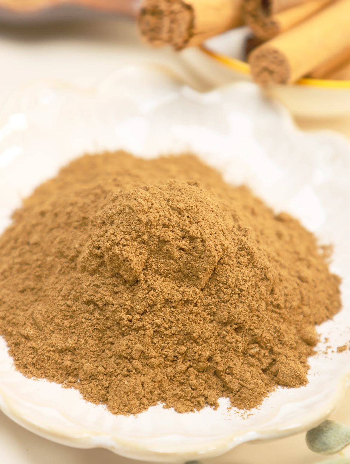 Fine golden-brown incense powder mounded on white plate with cinnamon sticks in background