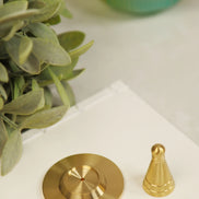 Golden copper incense cone maker with heavy circular base and pointed cone piece displayed on white marble surface