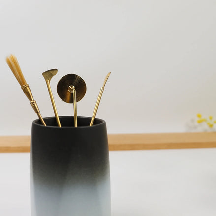 Black ceramic cylindrical holder containing gold brass wax sealing tools and implements on wooden surface