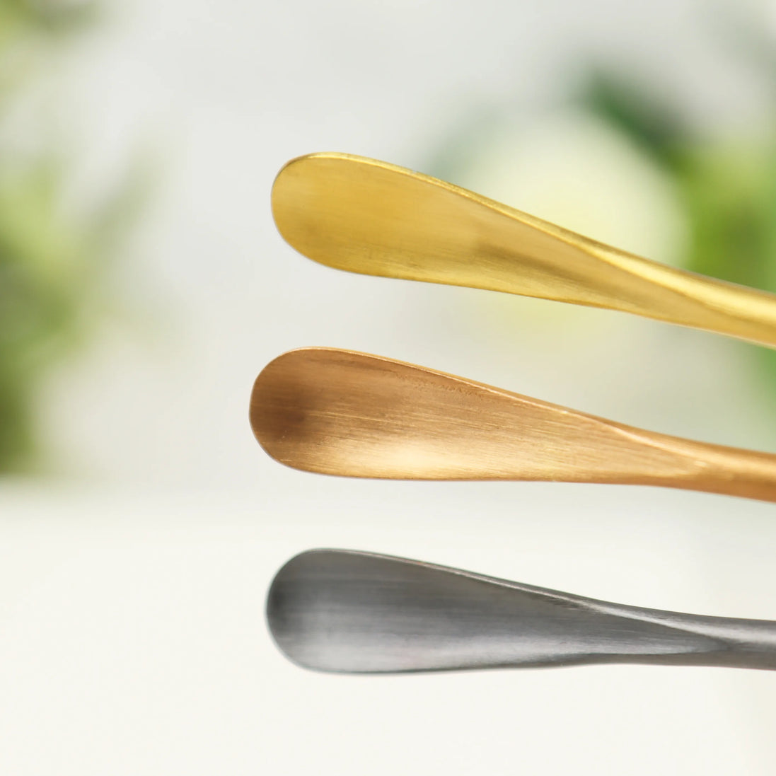 Three curved stainless steel incense scoops in rose gold, bronze, and silver finishes on white background