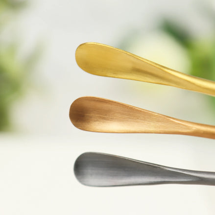 Three curved stainless steel incense scoops in rose gold, bronze, and silver finishes on white background