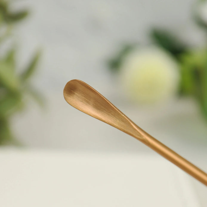 Rose gold stainless steel incense scoop with curved handle lying on white surface with blurred background