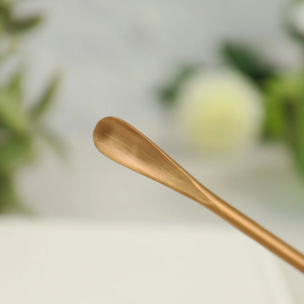 Rose gold stainless steel incense scoop with curved handle lying on white surface with blurred background