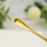 Gold-colored stainless steel incense scoop with curved bowl end resting on white surface with blurred background