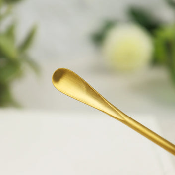 Gold-colored stainless steel incense scoop with curved bowl end resting on white surface with blurred background