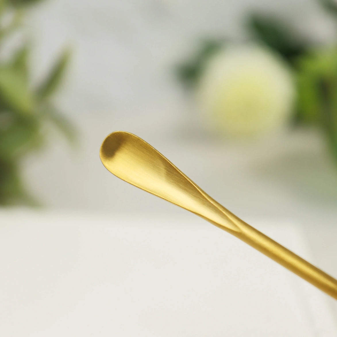 Gold-colored stainless steel incense scoop with curved bowl end resting on white surface with blurred background