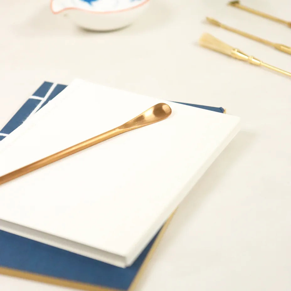 Rose gold stainless steel incense scoop lying on white paper with blue accents and shadows