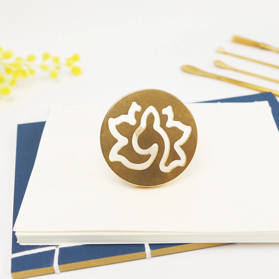 Round brass seal with organic lotus cutout design resting on white paper with navy and gold stationery items