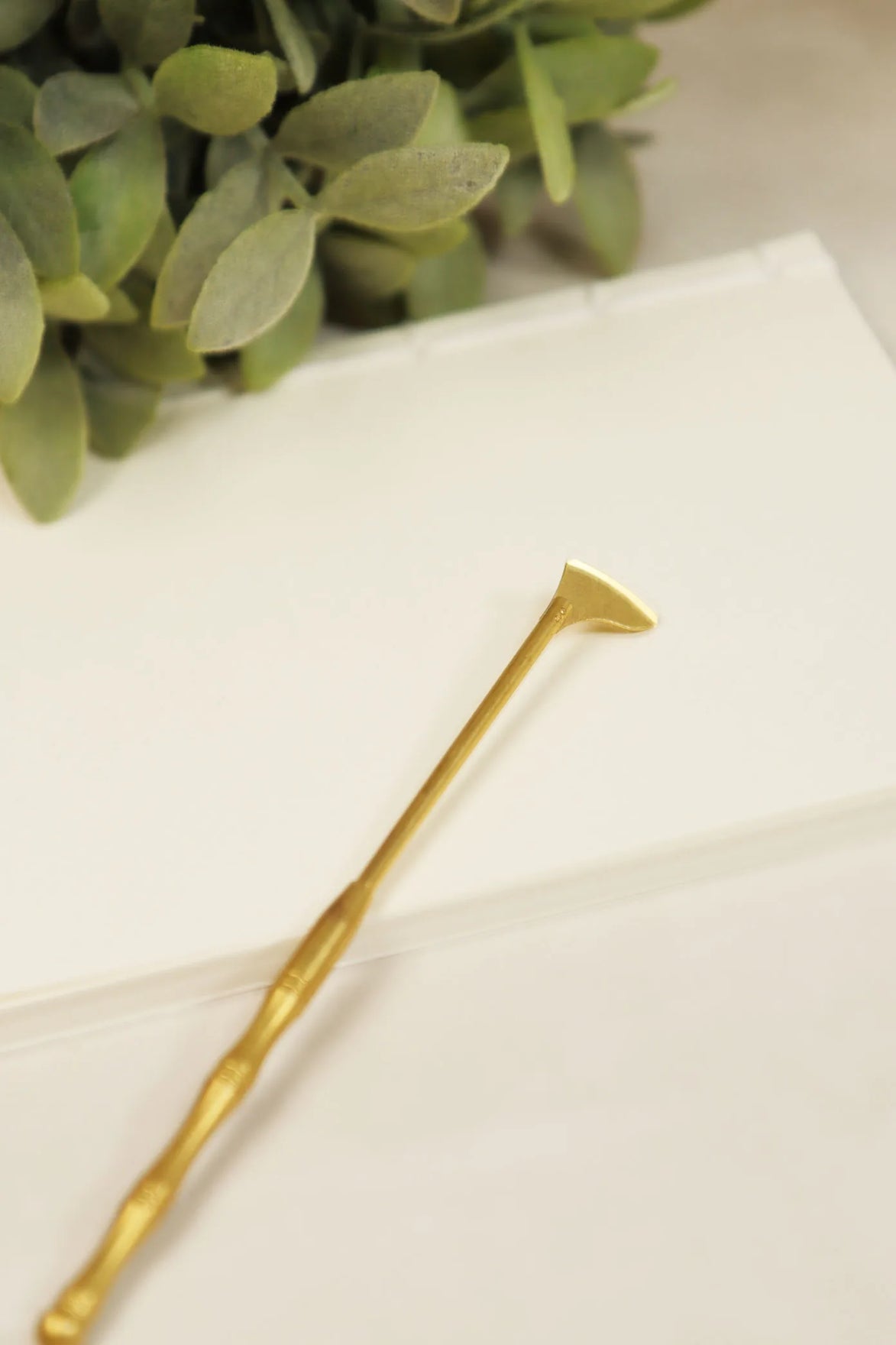 Golden brass spade tool with flat angled head and long handle on white marble surface with eucalyptus leaves