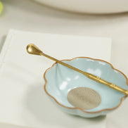 Brass incense spoon with long handle resting on scalloped mint green ceramic bowl with gold rim