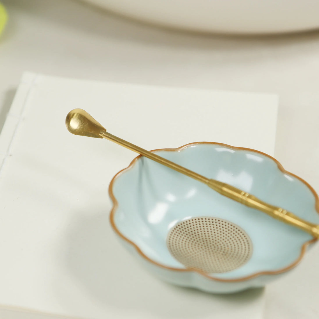 Brass incense spoon with long handle resting on scalloped mint green ceramic bowl with gold rim