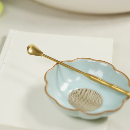 Brass incense spoon with long handle resting on scalloped mint green ceramic bowl with gold rim