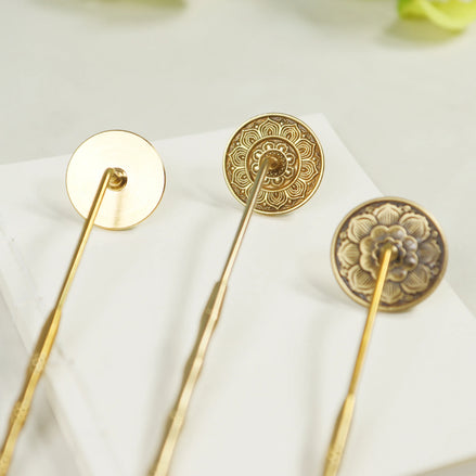 Three brass incense stamps with ornate lotus and mandala patterns on long handles arranged on white surface