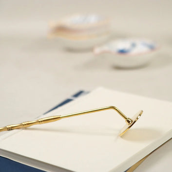 Gold brass incense sealing tool with curved handle resting on white surface for smoothing ash powder beds