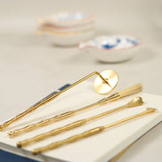 Four brass incense tools with gold finish including lotus stamp, brush, spade, spoon and scoop laid on white surface