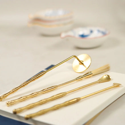 Four brass incense tools with gold finish including lotus stamp, brush, spade, spoon and scoop laid on white surface