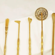 Five brass incense tools with ornate floral stamp, including brush, spade, scoop and spoon arranged on white surface