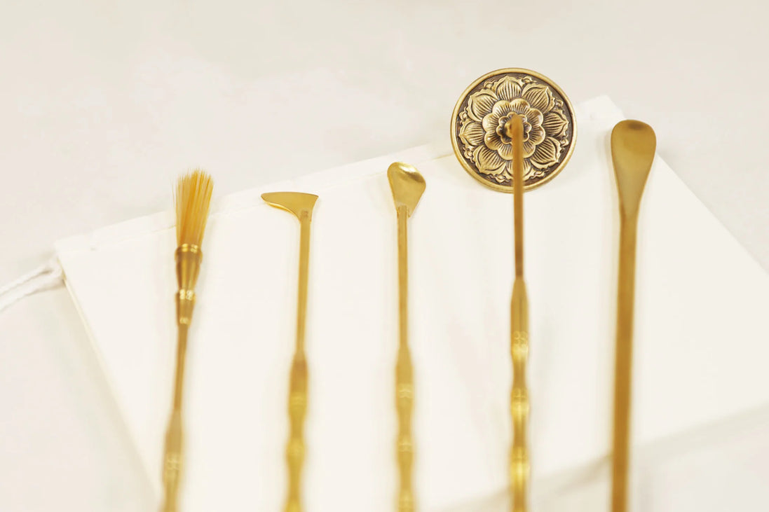 Five brass incense tools with ornate floral stamp, including brush, spade, scoop and spoon arranged on white surface
