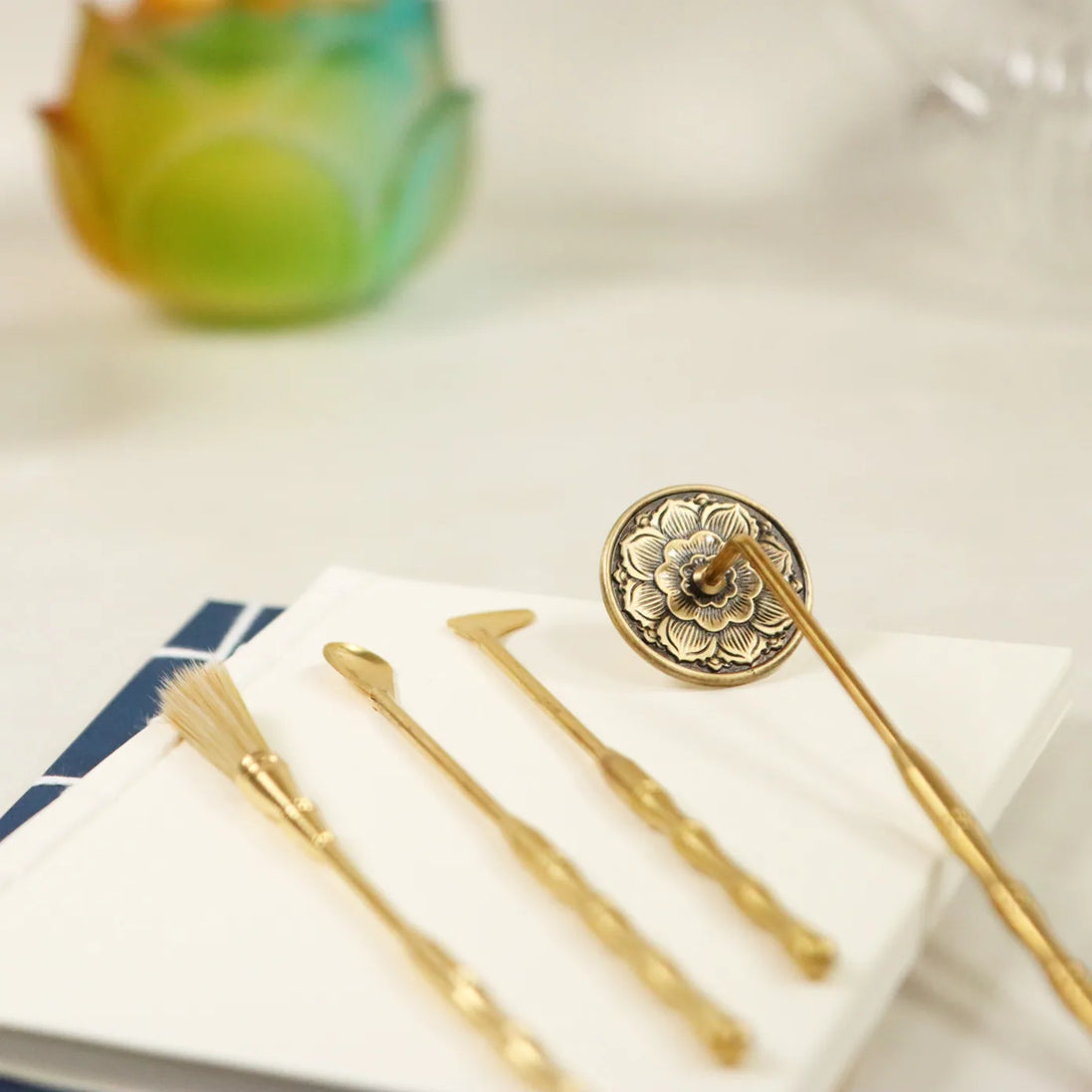 Four brass incense tools with ornate floral stamp displayed on white paper with colorful glass in background