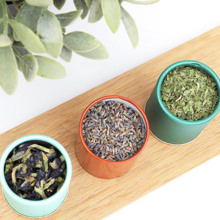Three colourful travel tins filled with loose leaf tea on bamboo tray