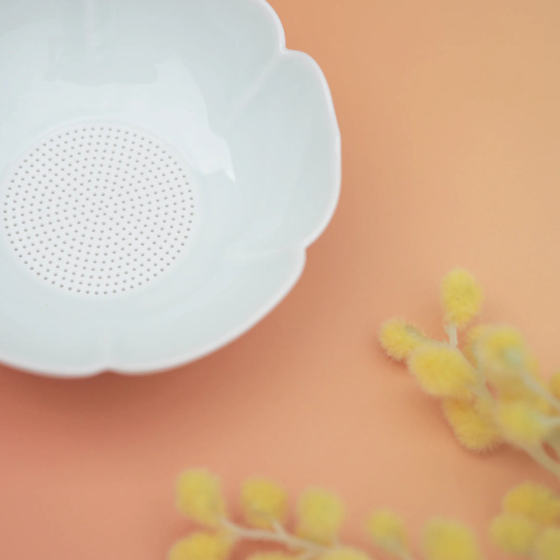 White flower-shaped tea strainer with fine mesh on coral background with yellow dried flowers